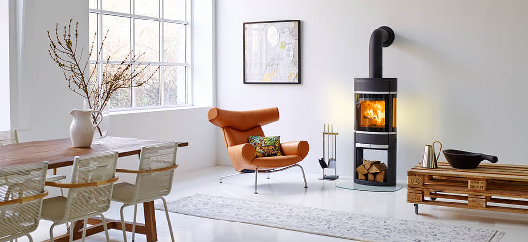 Svart oval vedovn Scan 68 i et hvitt rom med en designerstol i lyst skinn og et røft pallebord med hjul