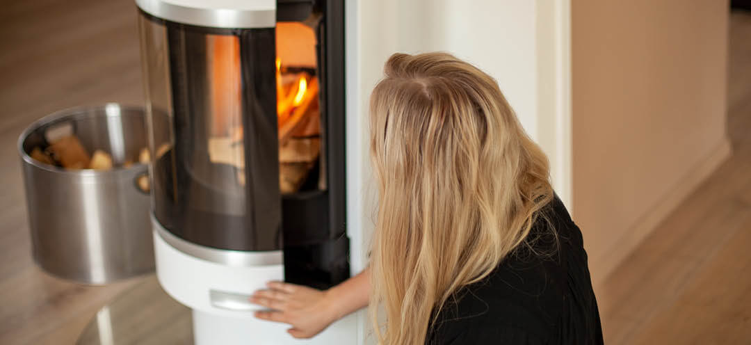 Blond kvinna sitter vid en vit vedspis Scan 83-2 och tänder brasan