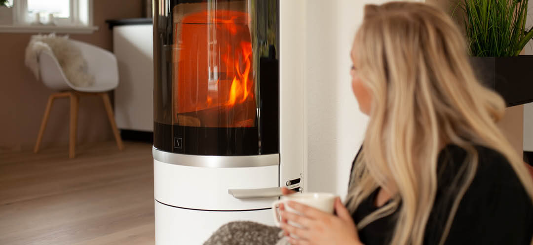 Blond kvinna sitter vid en vit vedspis Scan 83-2 och dricker te