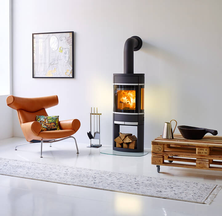 Svart öppen spis Scan 68 brinner i ett vitt rum med en cognacfärgad stol och ett pallbord bredvid