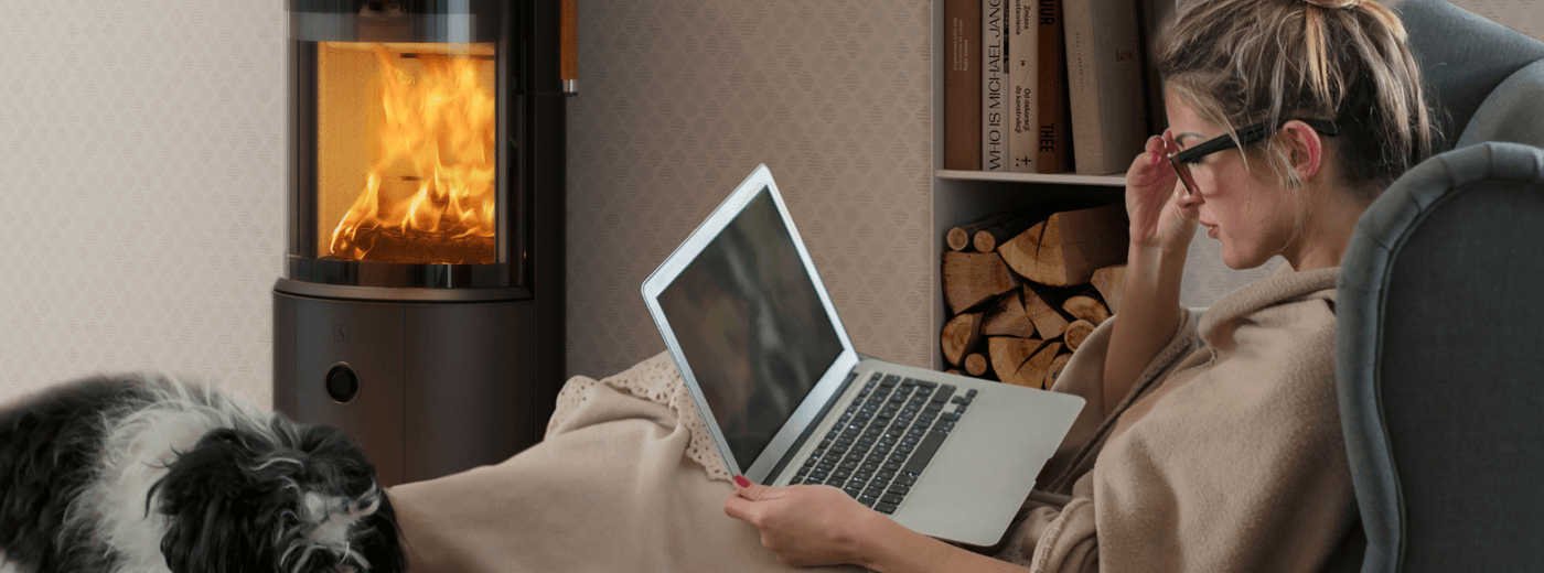 Kvinna sitter med laptop framför Scan 67 1600 med Zensoric, omgiven av böcker, ved och en hund – en smart kamin i ett modernt hem