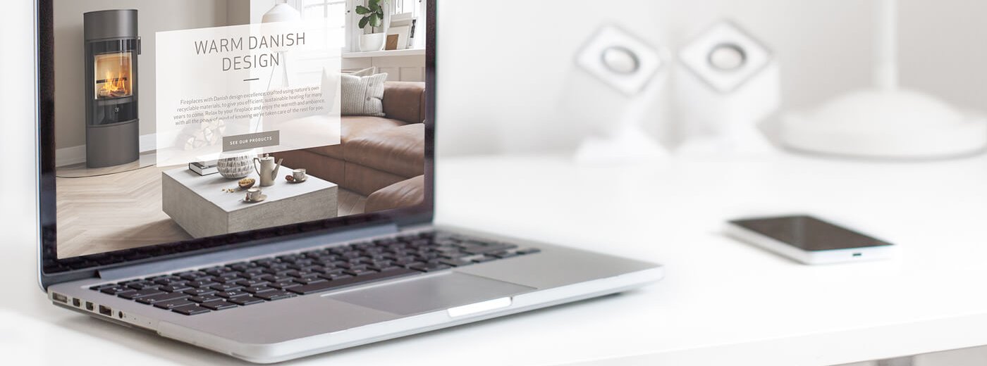 Bärbar dator som visar Scan Spis hemsida med modern vedkamin på skärmen, stående på ett vitt skrivbord bredvid mobiltelefon och högtalare